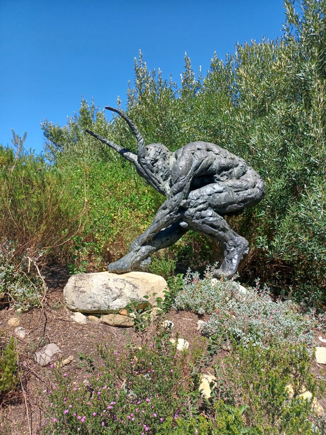 Crouching horned bronze figure among olive trees by Dylan Lewis